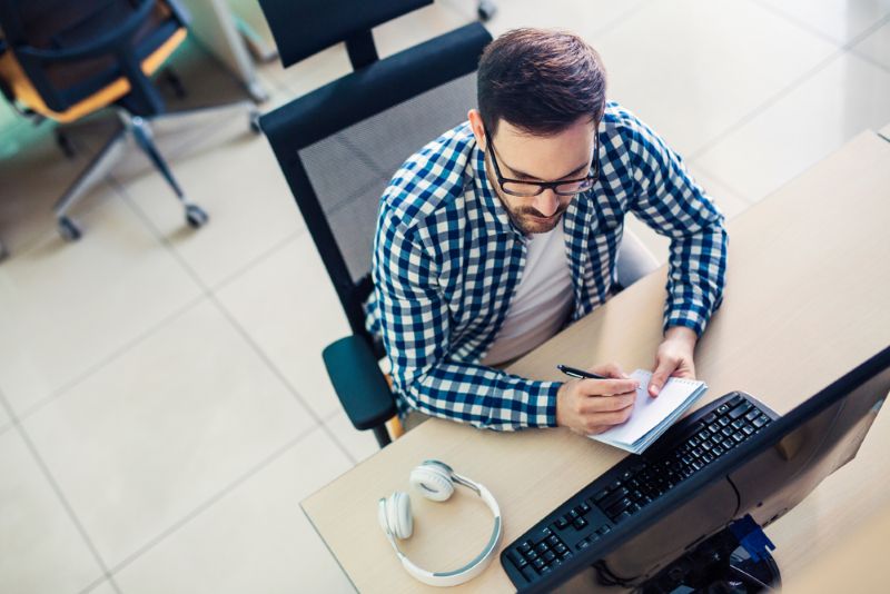 Male employee sitting at his desktop computer and taking notes on a notepad